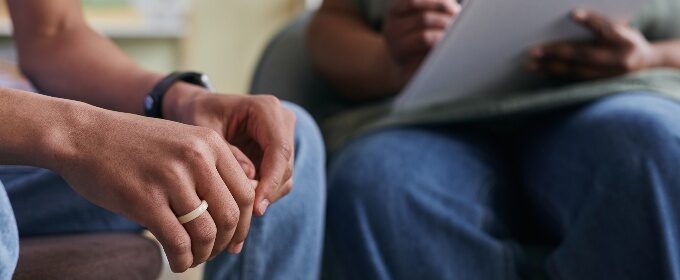 A close up image of a person's hands resting on their knees. Sitting next to them is somebody with a clipboard and pen.